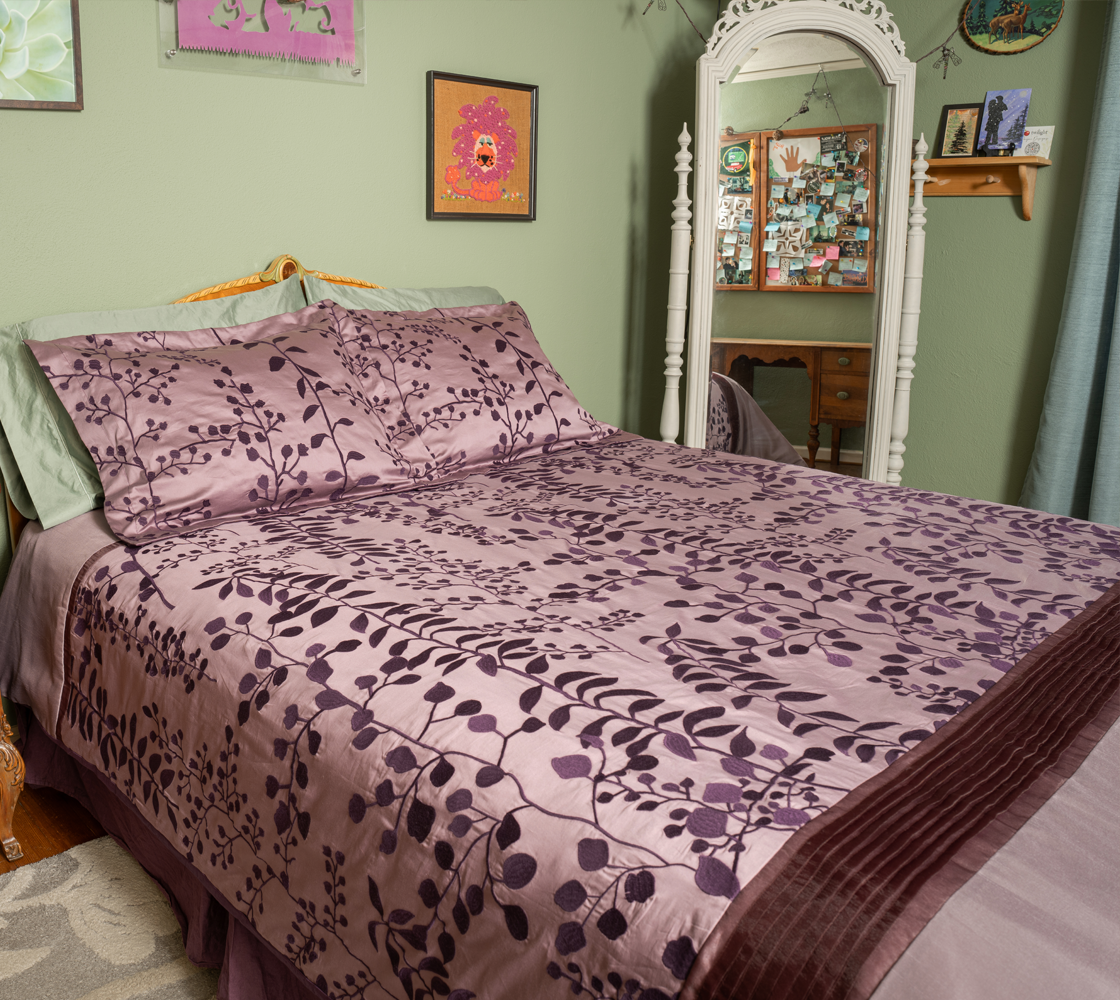 Bed with purple floral bedding in a room with a mirror and framed pictures on the wall.